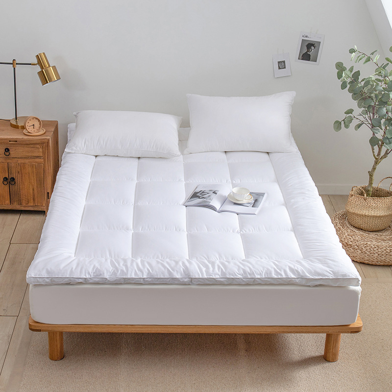 một chiếc giường màu trắng đặt topper trắng bên trên, trong phòng có sàn gỗ và một chậu cây, một kệ tủ, trên giường có một quyến sách và 2 cái gối trắng